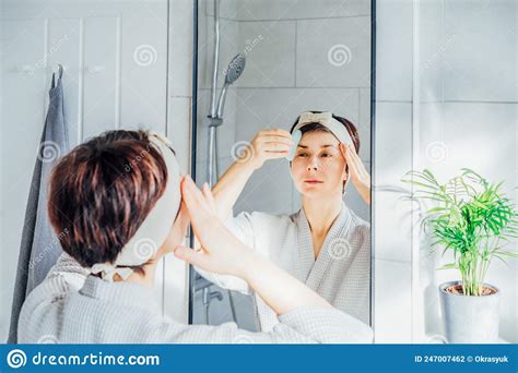 woman in bathrobe and hair band looking in the mirror and making face massage with gua sha