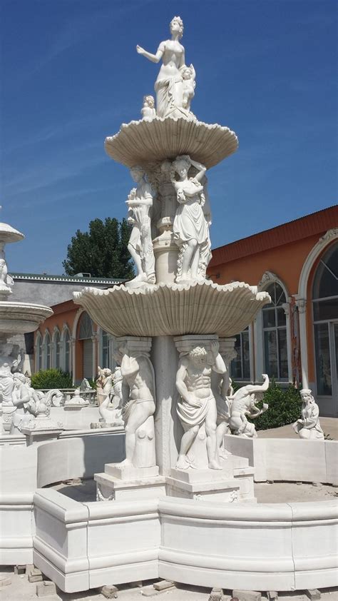 A White Fountain With Statues On It In Front Of A Building And Blue Sky Above