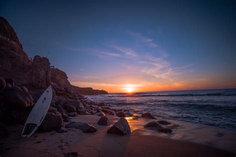 White Surfboard Leaning On Rock Near Water At Sunset Hd Wallpaper