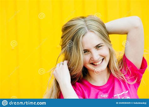 Beautiful Athletic Young Woman With Long Blond Hair In A Pink Shirt Posing And Smiling At Wall