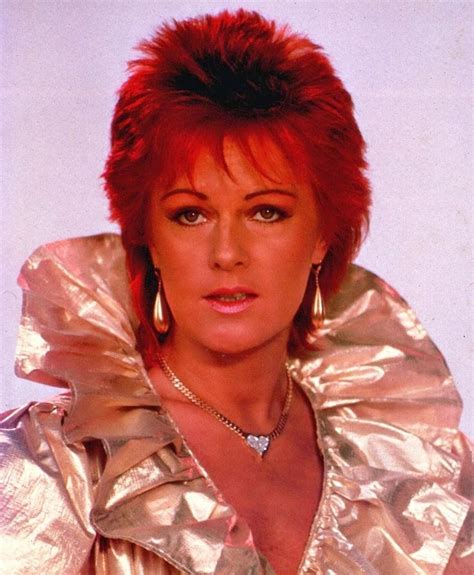 a close up of a person wearing a gold jacket and earrings with red hair in front of a white