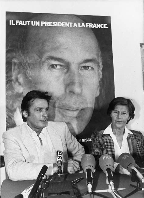 two people sitting at a table with microphones in front of them and a large poster behind them