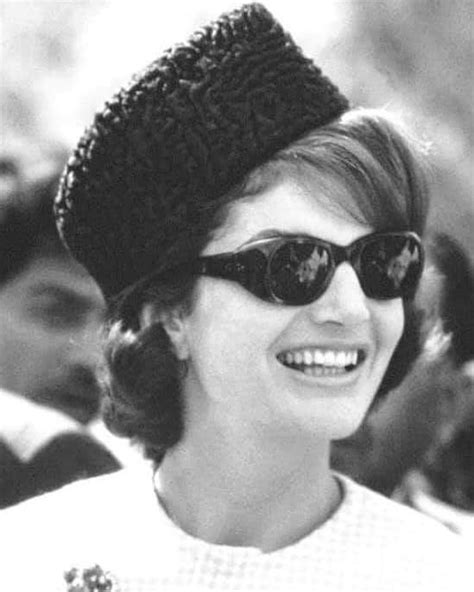 A Woman Wearing Sunglasses And A Knitted Hat Smiles At The Camera While Standing In Front Of A Crowd