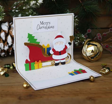 A Christmas Card With A Santa Clause On It Next To Gold Bells And Pine Cones