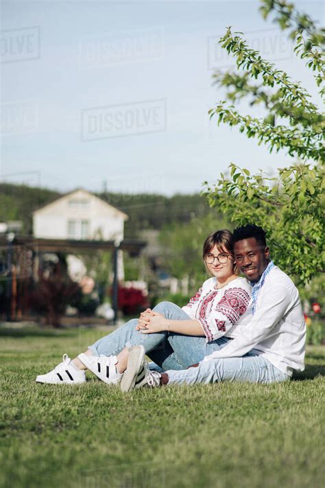 Interracial Couple Sits On Grass In Spring Garden Dressed In Ukrainian Traditional Ethnic