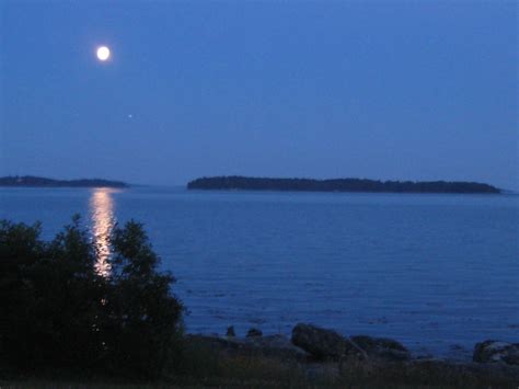The Full Moon Is Shining Brightly In The Sky Over The Water And Trees On The Shore