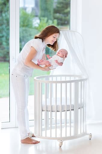 Young Mother Putting Newborn Baby To Sleep In White Crib Stock Photo