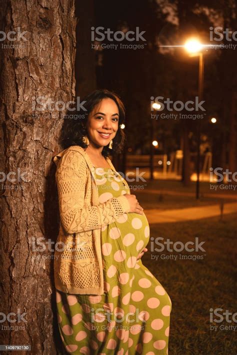Pregnant Latin Mom Leaning On A Tree In A Park At Night Looking At