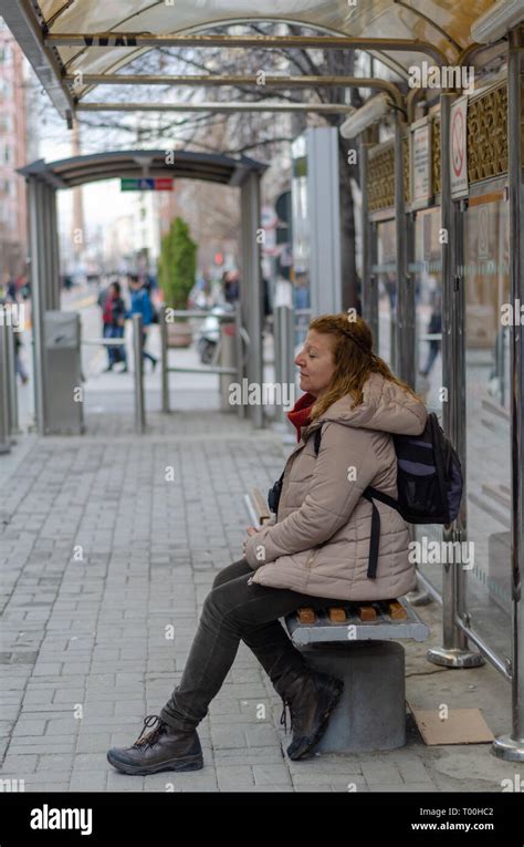 blonde woman is waiting for the tram at the stop she is tired and is sitting on the bench stock