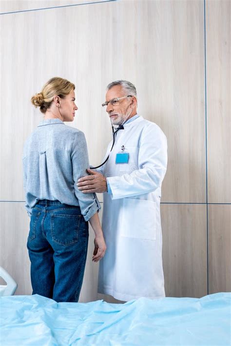 Confident Mature Male Doctor Examining Female Patient By Stethoscope