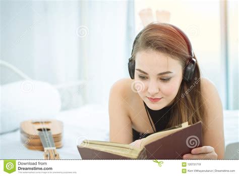 Girl Lying On Bed Reading Book And Listening To Music On Headphones