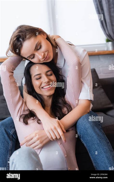 Smiling Lesbians With Closed Eyes Hugging In Living Room Stock Photo