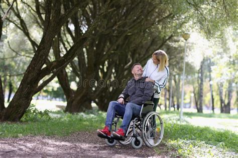 Woman Walking With Disabled Man In Wheelchair In Park Stock Photo