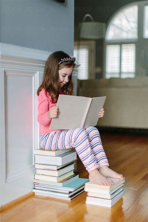 Cute Young Girl Sitting On A Stack Of Books Reading By Stocksy