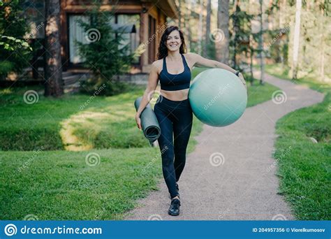 Outdoor Shot Of Happy Sporty Brunette Woman Walks On Path Near Forest Holds Rolled Up Karemat