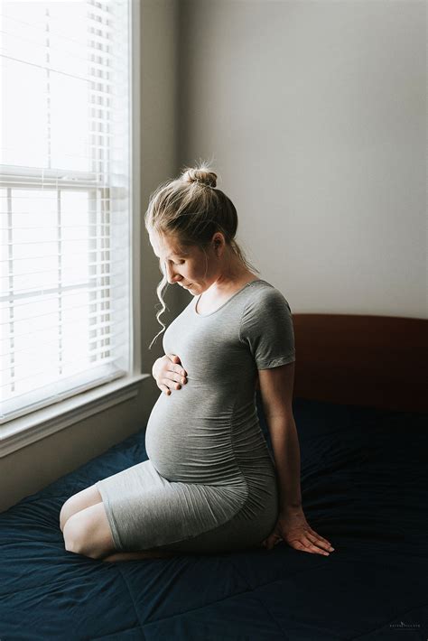 A Pregnant Woman Is Sitting On Her Bed Looking Out The Window While Holding Her Stomach