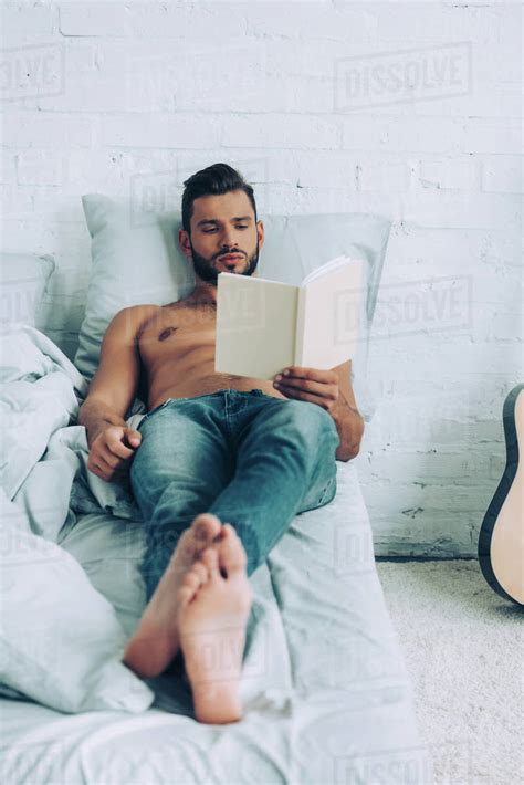 Focused Shirtless Muscular Man Reading Book During Morning Time In Bedroom At Home Stock Photo