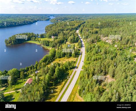 Aerial View Of Road Between Green Summer Forest And Blue Lake In