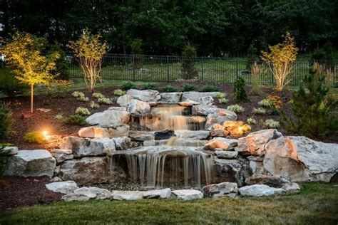 A Small Waterfall In The Middle Of A Garden With Lights On Its Sides