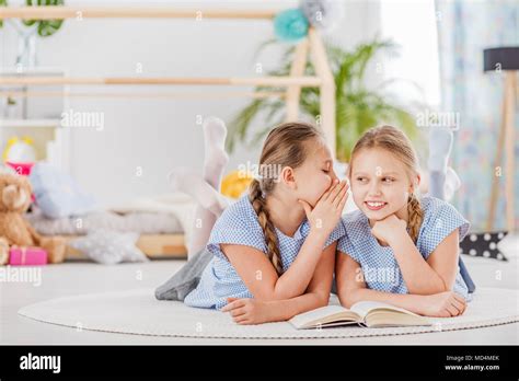 Mischievous Twin Sisters Whispering Secrets Lying On A White Rug In A
