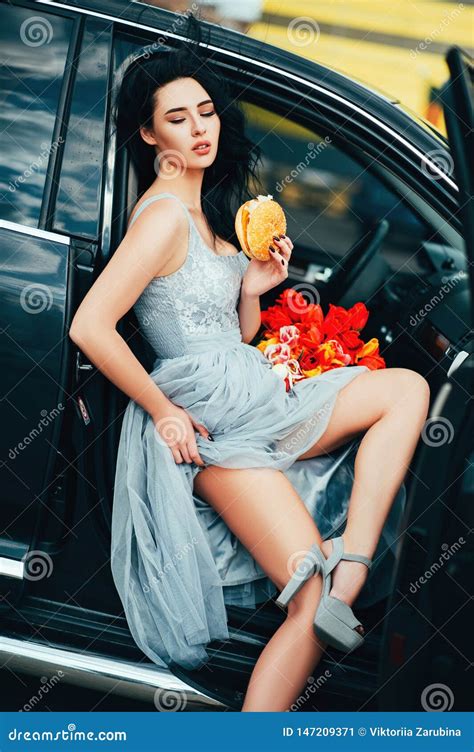 Portrait Of Elegance Sexual Woman In Dress With Flowers Sitting In Car And Eating Burger