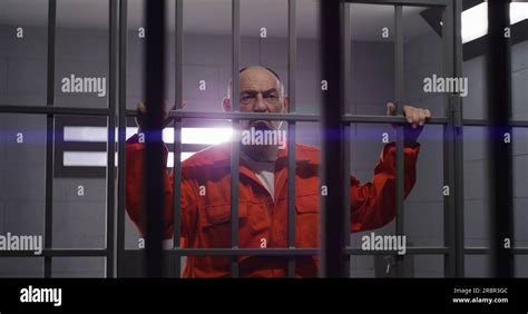 Elderly prisoner in orange uniform holds hands on metal bars. Criminal
