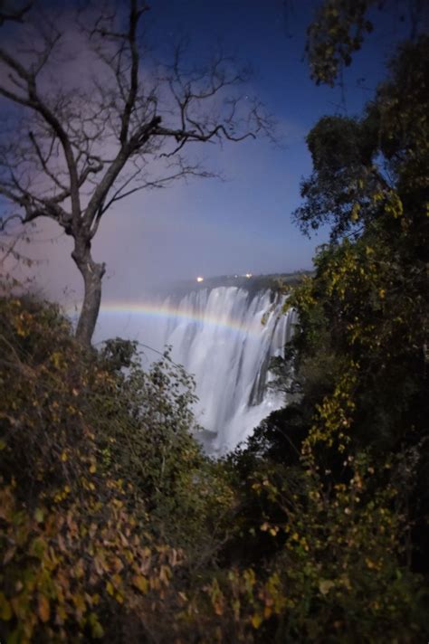 A Rainbow Shines In The Sky Over A Waterfall With Trees And Bushes