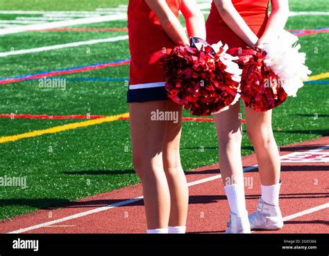 Rear View Of Two High School Cheerleaders Holding Their Pom Poms Behnd Their Back During A