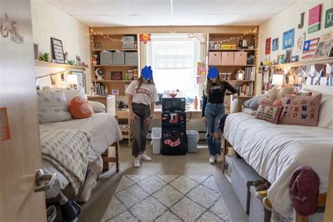 Two People Standing In A Dorm Room With Beds And Suitcases On The Floor