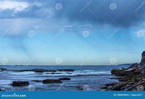 Coastal Rain Falling Over The Ocean Against Blue Sky With Seaside Rock
