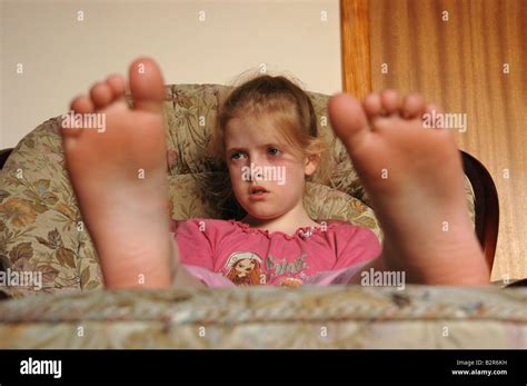 A Young Seated Girl Watches Tv With Her Bare Feet Resting On A Stool