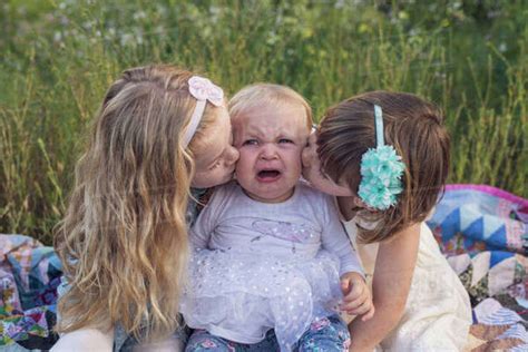 Girls Kissing Crying Sister While Sitting On Blanket In Park Stock