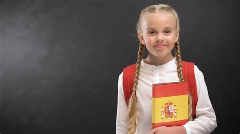 Smiling Schoolgirl Holding Spanish Language Book Against Blackboard