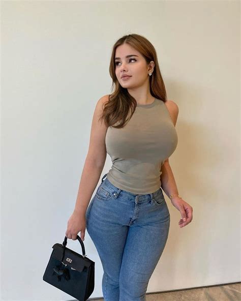 A Woman In High Rise Jeans Holding A Black Bag And Posing For The Camera With Her Right Hand On