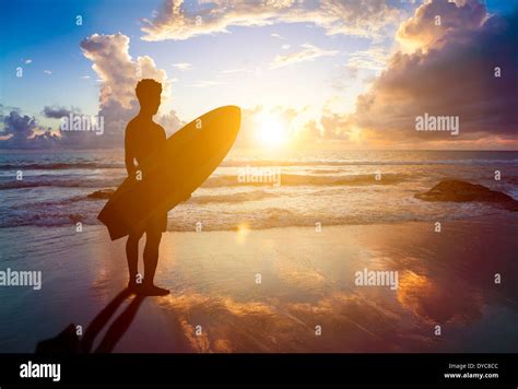 Man Standing On Beach At Sunrise Holding Surfboard Hi Res Stock