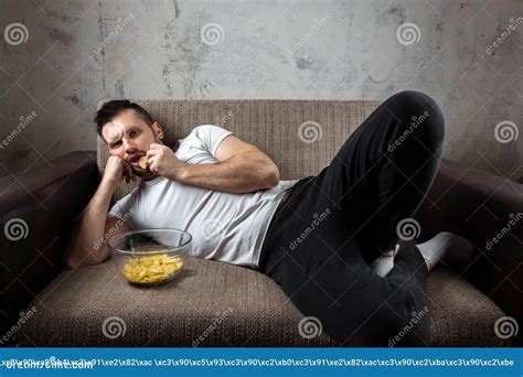 The Guy In The Shirt Is Lying On The Couch Eating Chips And Watching A