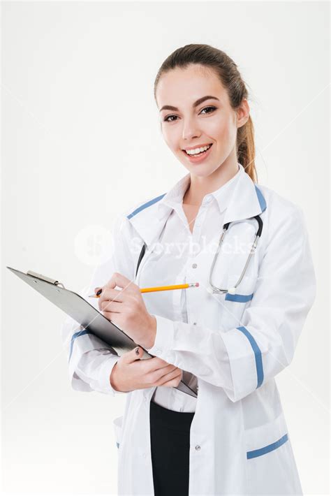 Cheerful Cute Young Woman Doctor Standing And Writing On Clipboard Over