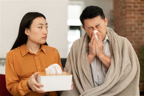 Sick Asian Man Wrapped In Blanket Sitting On Sofa And Sneezing Nose