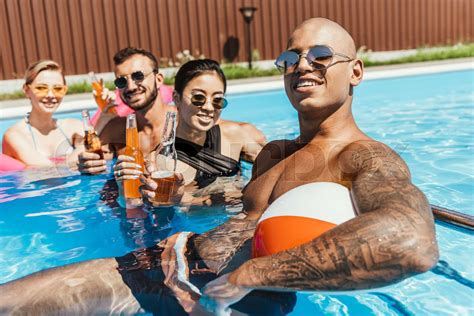 multiethnic friends with bottles of beer relaxing in water in swimming pool stock image