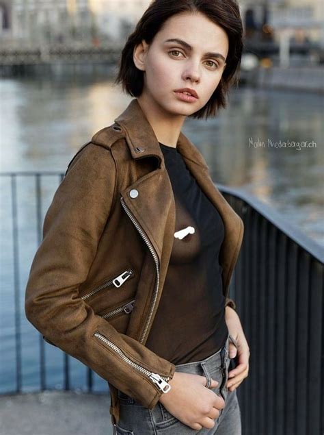 a woman standing next to a body of water wearing a brown leather jacket