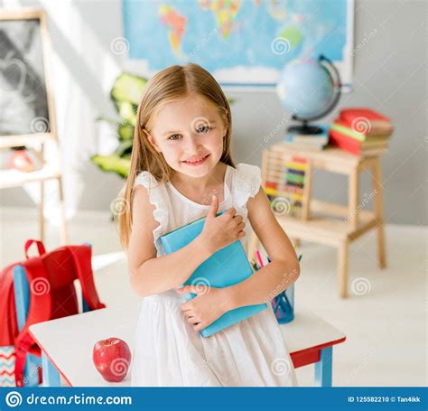 Little Smiling Blond Girl Holding Blue Book In The School Class Stock