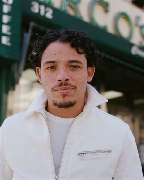 A Man With Curly Hair Wearing A White Jacket And Looking At The Camera