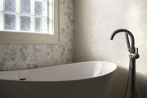 Luxurious bathroom nook features a marble slab accent wall framing a window covered in a gray grosgrain roman shade hung above a modern cast iron bathtub placed on marble floor tiles behind a round marble and brass accent table. Spa Bathroom with marble Hex Tile Accent Wall ...