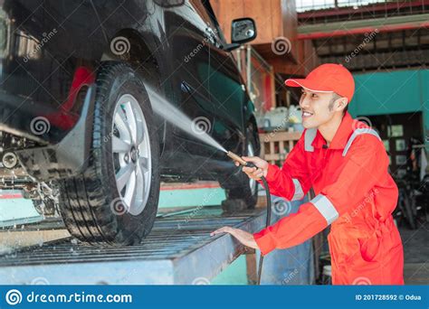 smiling asian male car cleaner wearing red uniform and hat is spraying water on a car stock