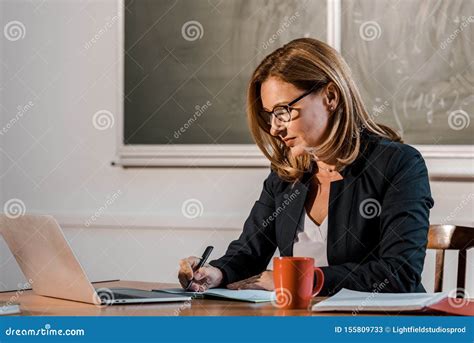 Female Teacher Sitting At Computer Desk And Writing In Notebook During Lesson Stock Image