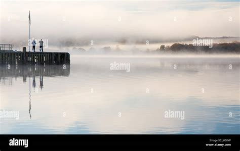 Two People Stand On Waterhead Pier In A Misty Windermere Lake At