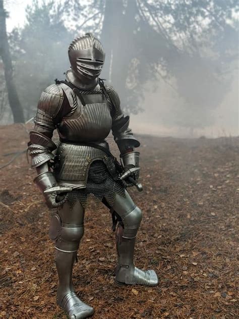 A Man Dressed In Armor Standing On Top Of A Dirt Field With Trees In