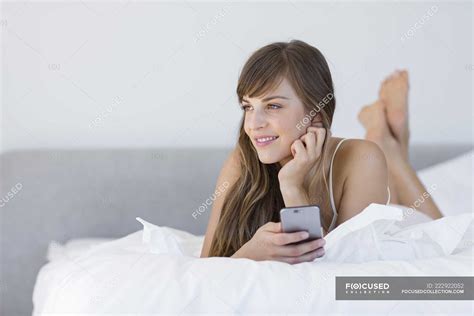 Smiling Young Woman Lying On Bed With Smartphone — Blanket Housing