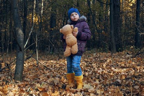 A Girl Child In Autumn Clothes Stands In The Forest On Dry Leaves And