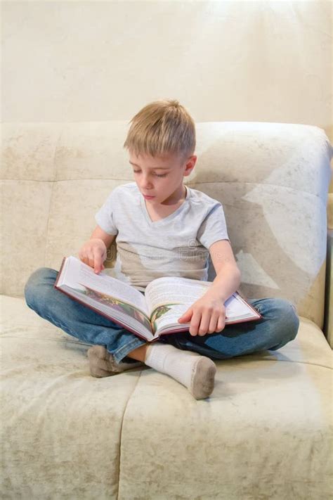 Little Boy Reads Book Sitting At Home On Beige Sofa And Is Very